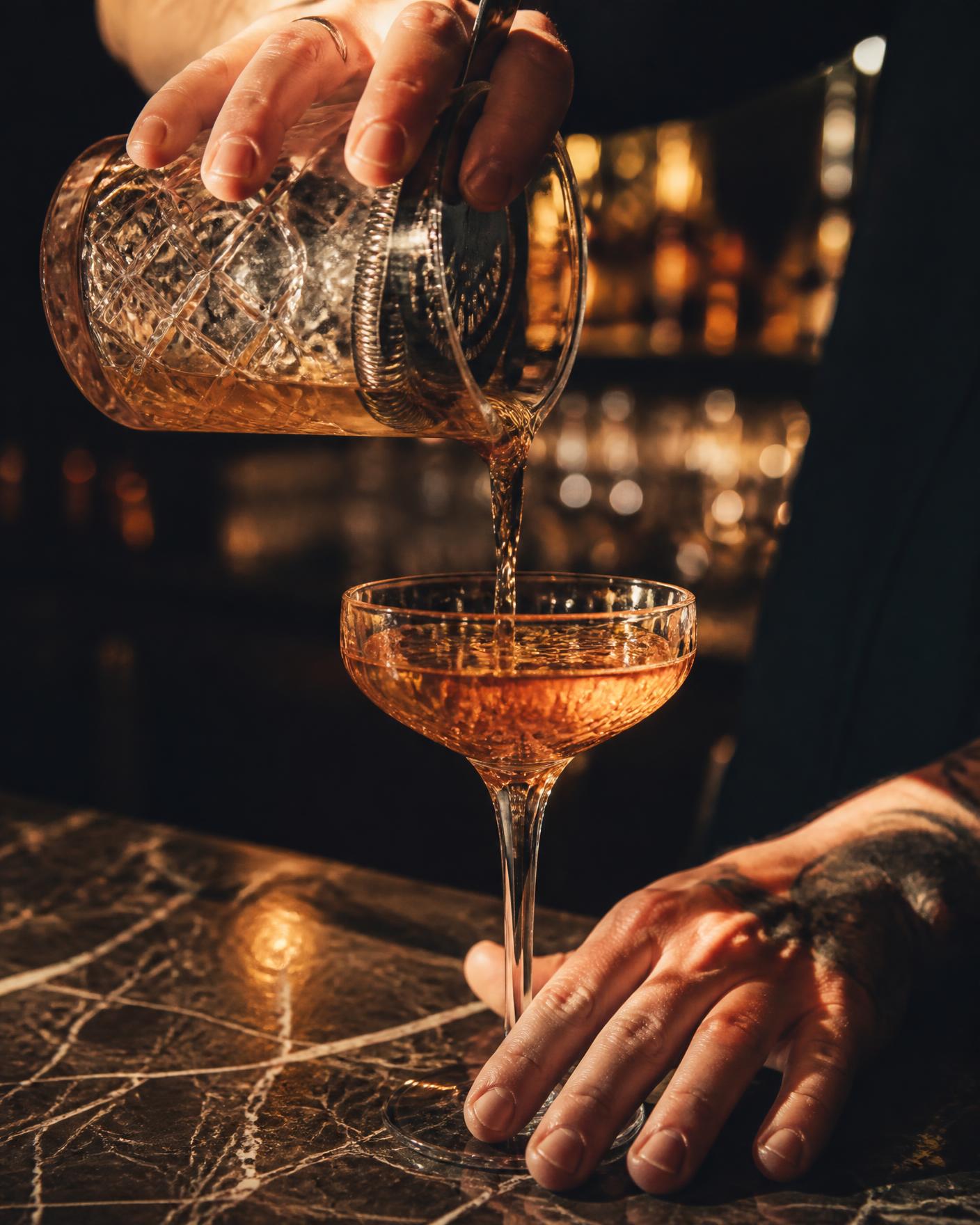Bartender pouring an amber cocktail into a coupe glass at a dark marble bar